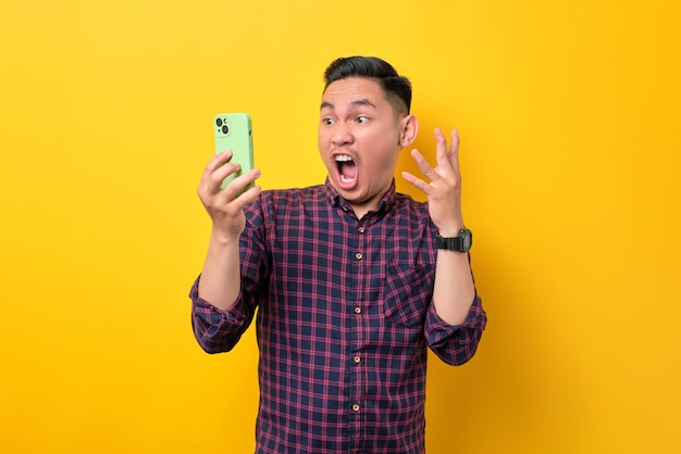 Удивленный молодой азиатский мужчина смотрит на экран смартфона и эмоционально реагирует на онлайн-новости, изолированные на желтом фоне