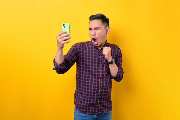 Surprised young Asian man looking at smartphone screen and raising hand with excitement isolated over yellow background