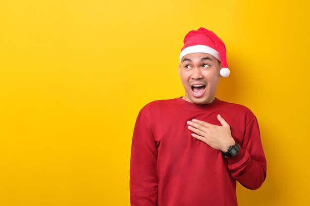 surprised young asian man in santa hat looking aside at copy space on yellow studio background celebration christmas holiday and new year concept