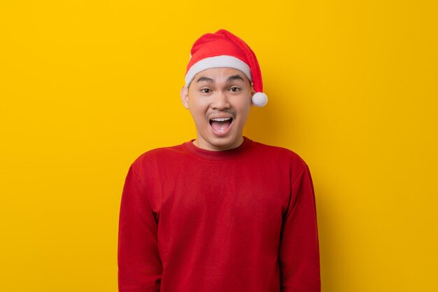 surprised young asian man in santa hat looking at camera with open mouth on yellow studio background celebration christmas holiday and new year concept