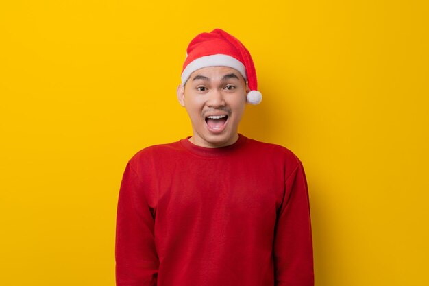 Surprised young asian man in santa hat looking at camera with open mouth on yellow studio background celebration christmas holiday and new year concept