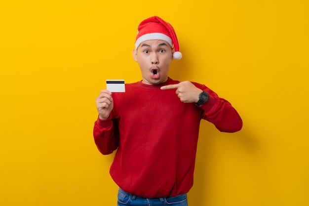 Surprised young Asian man in Santa hat pointing finger at credit card on yellow studio background celebration Christmas holiday and New Year concept