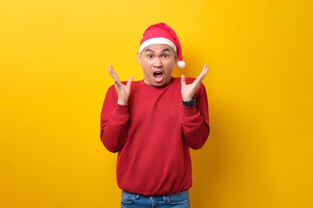 surprised young asian man in santa hat raising hands and looking at camera with excitement expression on yellow studio background celebration christmas holiday and new year concept