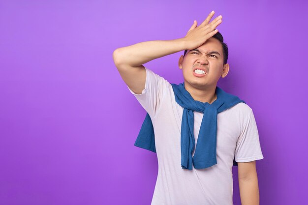 Surprised young Asian man wearing white tshirt put hand on face epic fail mistaken omg gesture isolated over purple background people lifestyle concept