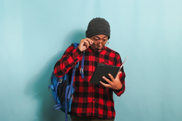 Surprised young Asian student in a beanie hat is reading a book with an astonished expression