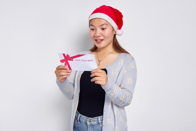 Surprised young asian woman 20s in hat christmas receives a\
gift certificate coupon voucher card isolated on white studio\
background merry christmas concept