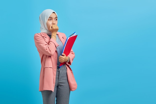 Surprised young asian woman holding folder and looking at empty\
spaceover blue background