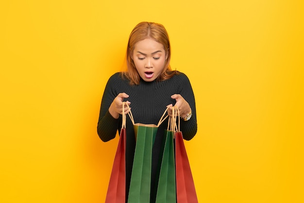 Surprised young Asian woman open shopping bags with open mouth isolated over yellow background