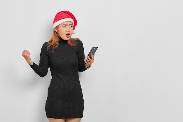 Surprised young Asian woman in Santa Claus hat holding mobile phone and looking away at copy space isolated over white background