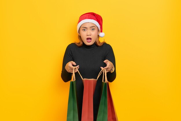 Surprised young Asian woman in Santa Claus hat open shopping bags with open mouth isolated over yellow background