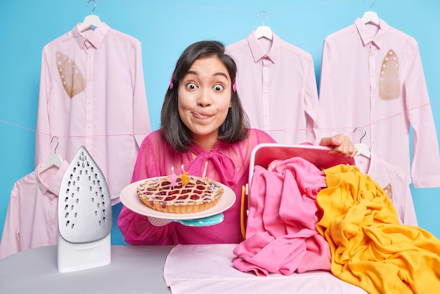 Surprised young asian woman stares impressed licks lips holds\
appetizing cherry cake stands near ironing board with pile of\
laundry against white shirts hanging on hangers over blue\
background