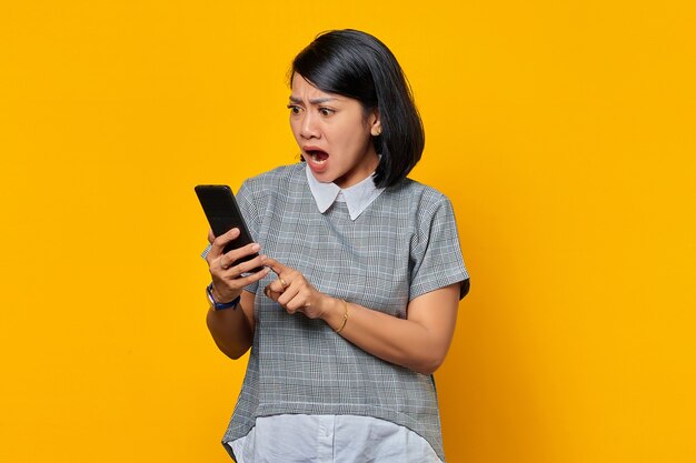 Удивленная молодая азиатская женщина с помощью мобильного телефона и глядя на экран смартфона на желтом фоне