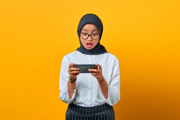 Surprised young Asian woman using mobile phone playing game on yellow background