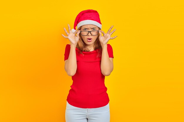 Фото Удивленная молодая азиатская женщина в рождественской шляпе в очках с открытым ртом на желтом фоне