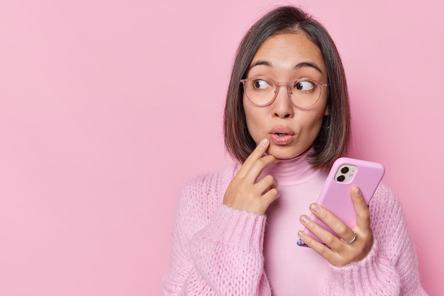 Surprised young asian woman with dark hair holds mobile phone\
and looks amazed away downloads new app reads shocking message\
wears warm jumper and big spectacles isolated over pink\
background