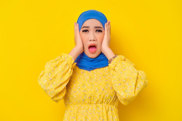 Surprised young beautiful asian muslim woman touching cheeks with open mouth isolated over yellow background