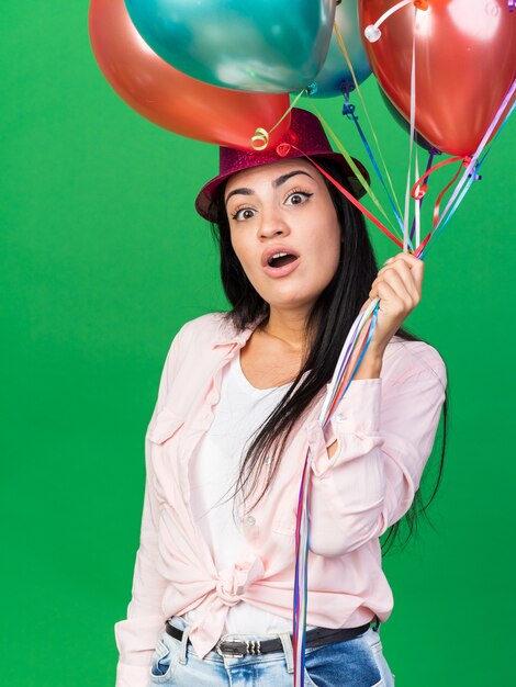 Giovane bella ragazza sorpresa che indossa il cappello del partito che tiene i palloni