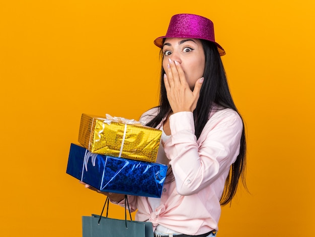 Surprised young beautiful girl wearing party hat holding gift bag with gift boxes covered mouth with hand isolated on orange wall