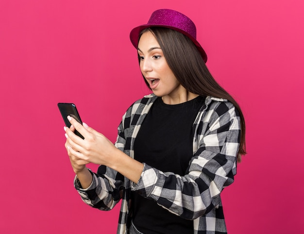 Surprised young beautiful girl wearing party hat holding and looking at phone 