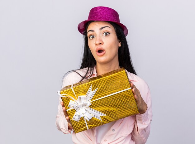 Surprised young beautiful girl wearing party hat holding out gift box at camera 