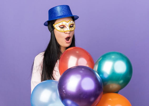 Photo surprised young beautiful girl wearing party hat and masquerade eye mask holding and looking at balloons isolated on blue wall