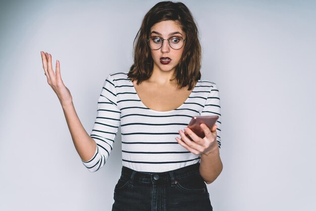 Surprised young beautiful woman browsing smartphone posing in studio