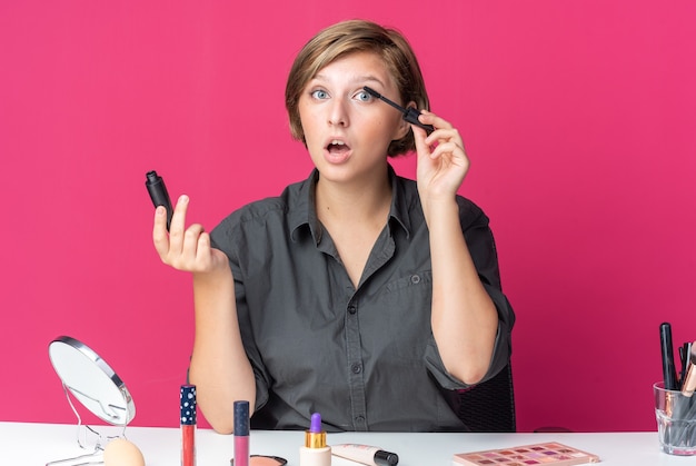 Surprised young beautiful woman sits at table with makeup tools applying mascara 