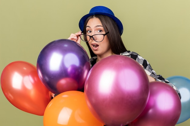 Surprised young beautiful woman wearing blue hat with glasses standing behind balloons isolated on olive green wall