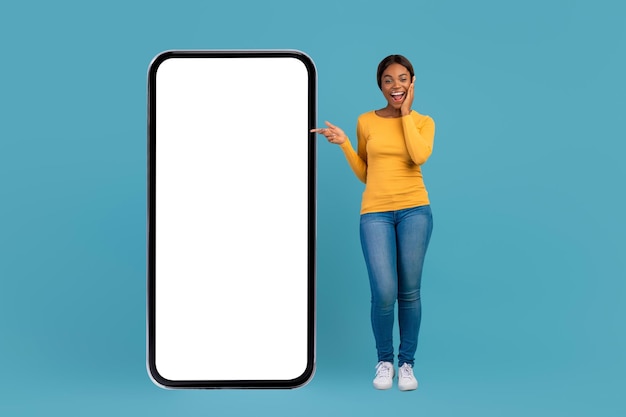 Surprised young black female pointing at big blank smartphone with blank screen