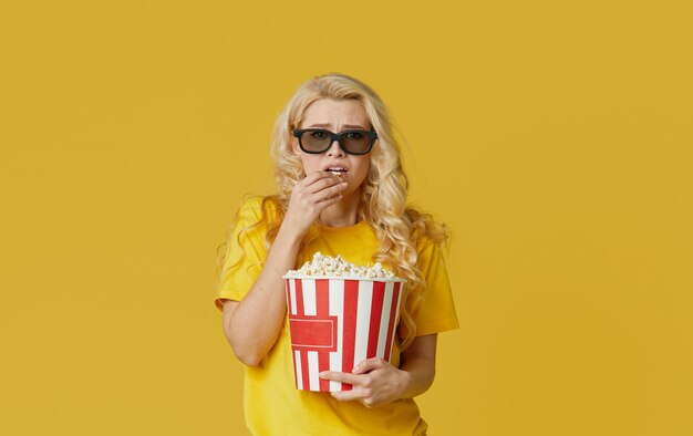 Surprised young blond woman in 3d glasses and yellow shirt eating popcorn, looks shocking movie at the cinema. Isolated