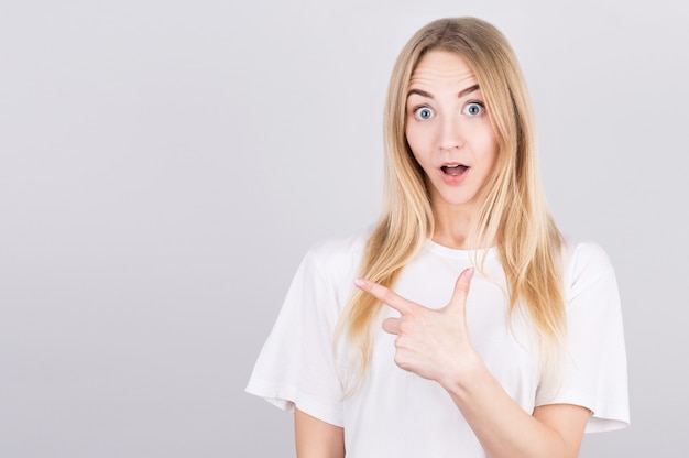 Surprised young blonde woman points her finger to the space to her left. Portrait of a surprised girl on a gray background.