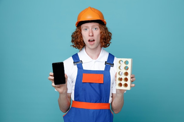 Sorpreso giovane costruttore uomo in uniforme tenendo il telefono con mattoni isolati su sfondo blu