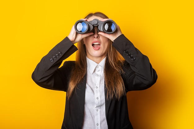 Surprised young business woman looking through binoculars on yellow.