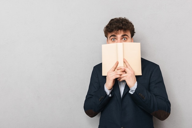 Surprised young businessman wearing suit standing isolated over gray, covering his face with a book