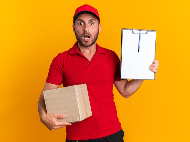 Surprised young caucasian delivery man in red uniform and cap holding cardboard box showing clipboard