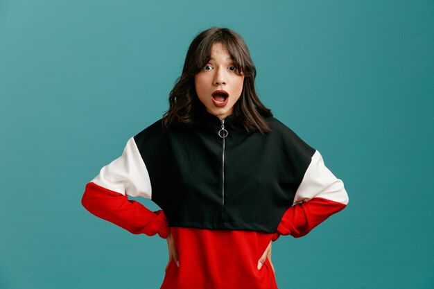 Surprised young caucasian woman keeping hands on waist looking at camera isolated on blue background