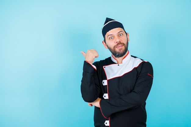 Surprised young chef is pointing back with thumb on blue background