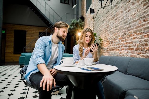 Giovani coppie sorprese che si siedono al tavolo in una caffetteria o in un caffè dell'hotel, guardando contenuti multimediali in uno smart phone