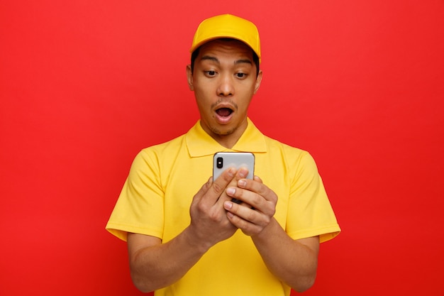 Surprised young delivery man wearing cap and uniform holding and looking at mobile phone 