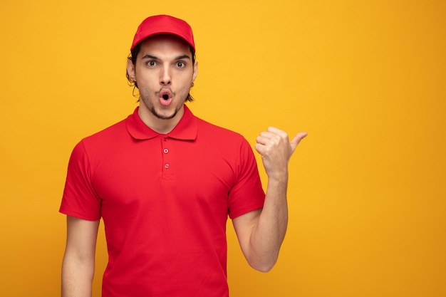 surprised young delivery man wearing uniform and cap looking at camera pointing to side isolated on yellow background with copy space