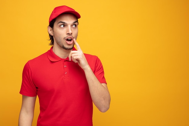 Giovane uomo di consegna sorpreso che indossa l'uniforme e il cappuccio che tocca il viso con il dito guardando il lato isolato su sfondo giallo con spazio per la copia