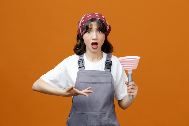 Sorpresa giovane donna addetta alle pulizie che indossa uniforme e bandana che mostra lo stantuffo che punta verso di esso con la mano che guarda la fotocamera isolata su sfondo arancione