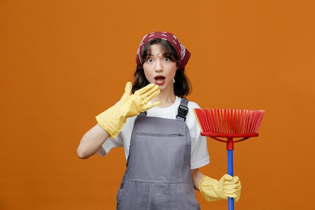 Surprised young female cleaner wearing uniform rubber gloves and bandana holding squeegee mop touching chin with hand looking at camera isolated on orange background