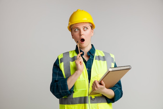 Giovane operaio edile femminile sorpreso che indossa il casco di sicurezza e la maglia di sicurezza che tiene il blocco note che tocca il fronte con la matita che esamina il lato