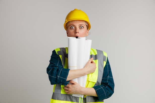 Surprised young female construction worker wearing safety helmet and safety vest hugging papers 