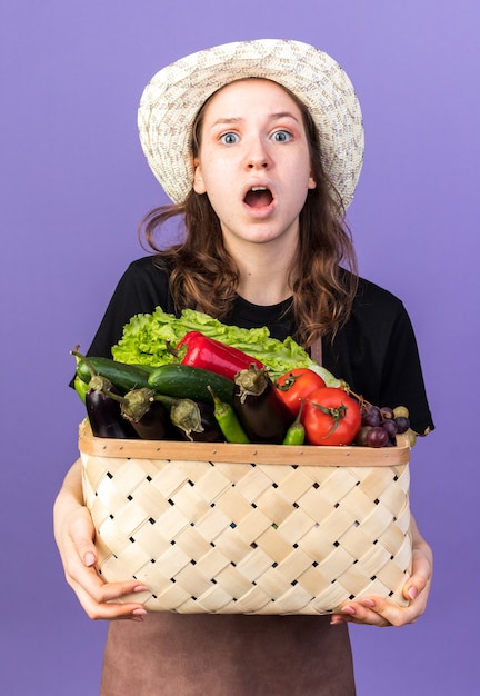 Surprised young female gardener wearing gardening hat holding vegetable basket 