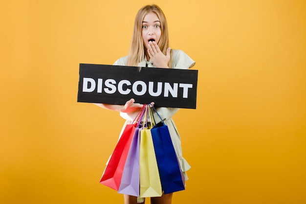 Photo surprised young girl has discount sign with colorful shopping bags isolated over yellow