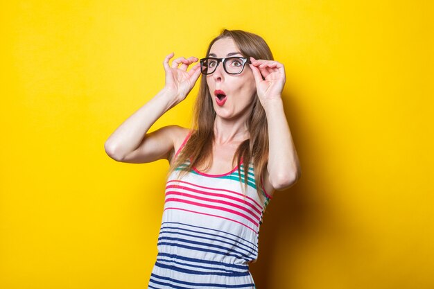 Surprised young girl holding glasses in a striped dress on a yellow background