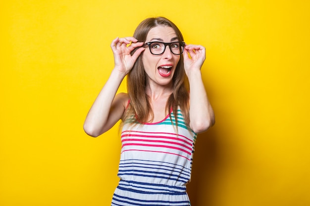 Surprised young girl holding glasses in a striped dress on a yellow background