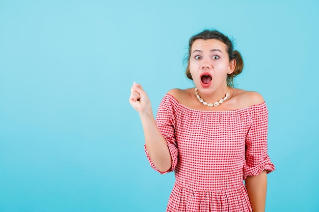 Surprised young girl is looking at camera by raising up her fist on blue background